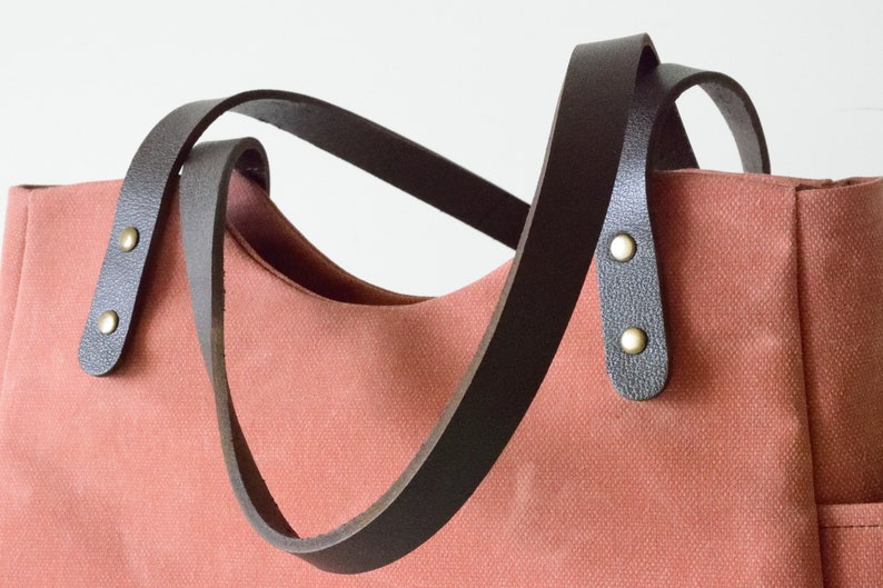 K&ouml;nnte beinhalten: Korallfarbene Tragetasche mit dunkelbraunen Ledergriffen. Die Griffe sind mit messingfarbenen Nieten am Beutel befestigt. Die Tasche hat ein schlichtes, klassisches Design, das f&uuml;r den t&auml;glichen Gebrauch geeignet ist.