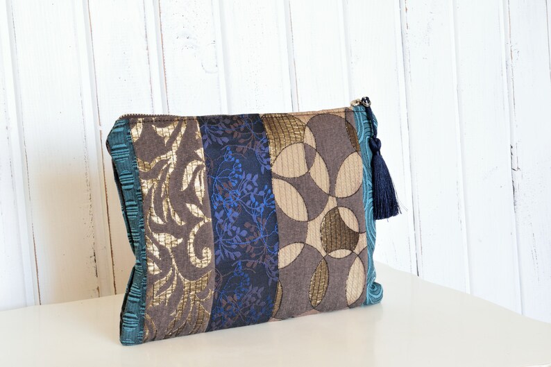 May include: A brown, blue and gold patchwork clutch with a blue tassel zipper pull. The clutch is made from a variety of fabrics with different patterns.