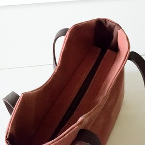 K&ouml;nnte beinhalten: Eine handliche, rosafarbene Tragetasche mit dunkelbraunen Griffen. Die Innenseite der Tasche ist in einem dunkleren Braunton gehalten. Ein schwarzer Rei&szlig;verschluss verl&auml;uft &uuml;ber die Oberseite der Tasche. Die Tasche ist aus robustem Material gefertigt.