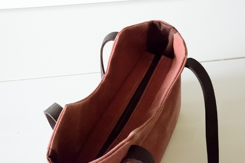 K&ouml;nnte beinhalten: Eine handliche, rosafarbene Tragetasche mit dunkelbraunen Griffen. Die Innenseite der Tasche ist in einem dunkleren Braunton gehalten. Ein schwarzer Rei&szlig;verschluss verl&auml;uft &uuml;ber die Oberseite der Tasche. Die Tasche ist aus robustem Material gefertigt.