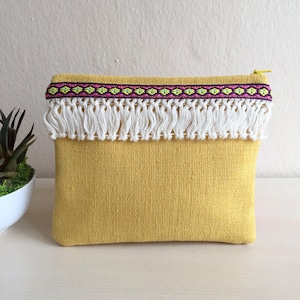 May include: A yellow fabric clutch with a white fringe trim and a colorful geometric pattern.