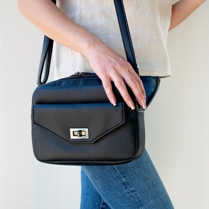 May include: A black leather crossbody bag with a silver clasp and a flap pocket. The bag is worn over the shoulder of a person wearing blue jeans.