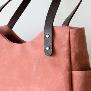 K&ouml;nnte beinhalten: Eine korallenfarbene Tragetasche mit dunkelbraunen Ledergriffen. Die Tasche hat eine geschwungene Oberseite und eine kleine Tasche an der Seite. Die Griffe sind mit messingfarbenen Nieten befestigt. Die Tasche besteht aus robustem Canvas-Material.