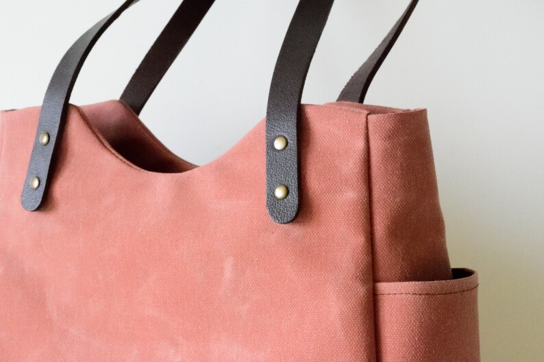 K&ouml;nnte beinhalten: Eine korallenfarbene Tragetasche mit dunkelbraunen Ledergriffen. Die Tasche hat eine geschwungene Oberseite und eine kleine Tasche an der Seite. Die Griffe sind mit messingfarbenen Nieten befestigt. Die Tasche besteht aus robustem Canvas-Material.