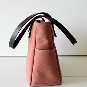 K&ouml;nnte beinhalten: Eine altrosa Tragetasche mit dunkelbraunen Ledergriffen. Die Tasche hat eine rechteckige Form mit einer einzelnen Au&szlig;entasche. Die Griffe sind gebogen und oben an der Tasche befestigt. Die Tasche ist aus robustem Canvas-Material.