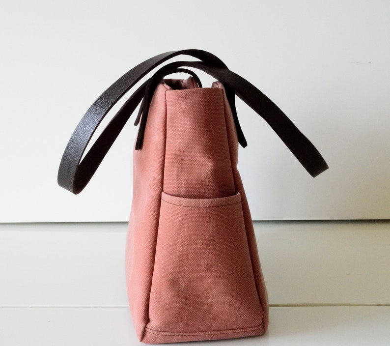 K&ouml;nnte beinhalten: Eine altrosa Tragetasche mit dunkelbraunen Ledergriffen. Die Tasche hat eine rechteckige Form mit einer einzelnen Au&szlig;entasche. Die Griffe sind gebogen und oben an der Tasche befestigt. Die Tasche ist aus robustem Canvas-Material.