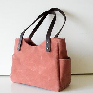 K&ouml;nnte beinhalten: Eine korallenfarbene Tragetasche mit dunkelbraunen Ledergriffen und Akzenten. Die Tasche hat zwei Au&szlig;entaschen und besteht aus robustem Canvas-Material. Die Griffe sind mit messingfarbenen Nieten befestigt.
