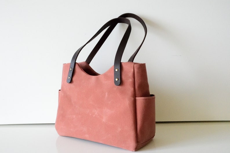 K&ouml;nnte beinhalten: Eine korallenfarbene Tragetasche mit dunkelbraunen Ledergriffen und Akzenten. Die Tasche hat zwei Au&szlig;entaschen und besteht aus robustem Canvas-Material. Die Griffe sind mit messingfarbenen Nieten befestigt.