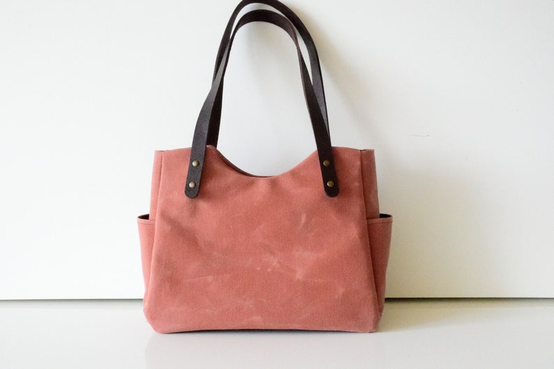 K&ouml;nnte beinhalten: Eine staubrosa Tragetasche mit dunkelbraunen Ledergriffen und Akzenten. Die Tasche hat zwei Au&szlig;entaschen und besteht aus einem robusten Canvas-Material. Die Griffe sind mit messingfarbenen Nieten befestigt.
