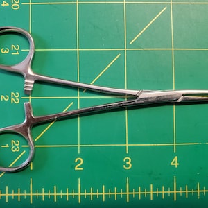 May include: A pair of silver hemostats, a type of surgical clamp, on a green cutting mat with yellow markings.