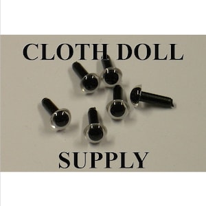May include: A close-up of black plastic safety eyes for crafts. The eyes are about 1/2 inch in diameter and have a clear plastic ring around the black pupil. The eyes are scattered on a white surface.  The text "CLOTH DOLL SUPPLY" is printed in black letters on the white surface.