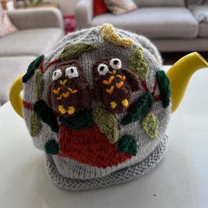 May include: A grey knitted teapot cosy with two brown owls and green leaves. The cosy is on a yellow teapot.
