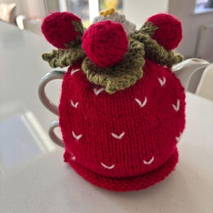 May include: A red knitted strawberry-shaped tea cosy with white seed details and green leaves. The cosy is topped with three red knitted strawberries and a white and yellow flower. Designed to fit a standard teapot.