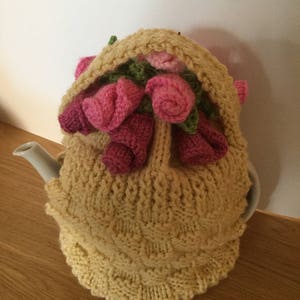 Può includere: Un copri-teiera in maglia di colore giallo chiaro con un motivo di rose rosa che spuntano dalla parte superiore.