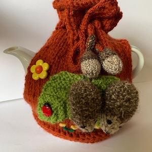 以下が含まれることがあります： オレンジ、グリーン、ブラウンのニットのティーポットカバーで、花とテントウムシのデザインが施されています。2匹の茶色のニットのネズミが、緑のニットの葉っぱの後ろから顔を出しています。