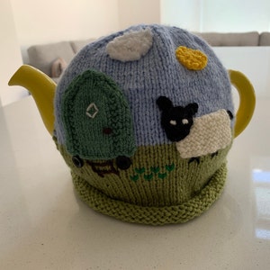 Op de afbeelding: Een gebreide theepotwarmer met een blauwe lucht, witte wolken, een gele zon, een groen huis en een wit schaap met een zwart gezicht. De warmer is groen aan de onderkant en heeft een gele theepottuit.