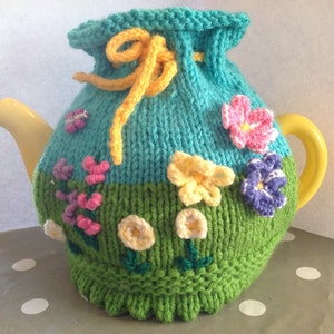 Könnte beinhalten: Eine gestrickte Teekannenhülle mit einem grünen und blauen Design mit Blumen und einem gelben Band. Die Hülle befindet sich auf einer gelben Teekanne.