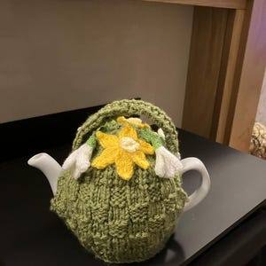 May include: A white ceramic teapot with a green knitted cozy. The cozy features a yellow flower and white flower accents. The teapot is on a black surface.