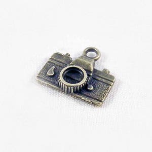 May include: A small, antique gold-toned camera charm with a detailed design. The charm has a loop at the top for attaching to a necklace or bracelet.