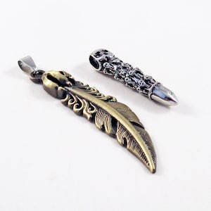 Könnte beinhalten: Zwei silberne und goldene Anhänger. Ein Anhänger ist eine Feder mit komplizierten Details. Der andere Anhänger ist ein kugelförmiges Design mit einem Schädel oben.