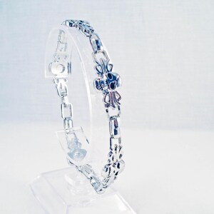 May include: A silver bracelet with a decorative, ornate design. The bracelet is on a white display stand.