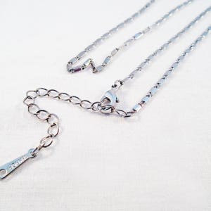 May include: A silver-colored necklace with a delicate, rectangular link chain. The necklace has an adjustable chain with a lobster clasp closure. The jewelry is displayed on a white surface.