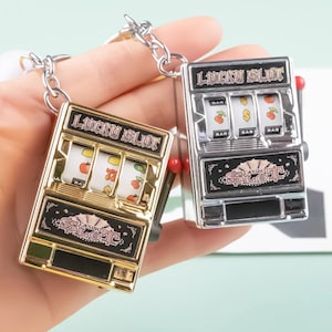 Peut inclure: Deux porte-clés en or et argent en forme de machines à sous avec les mots "Lucky Slot" et "Casino Slot" dessus. Les machines à sous ont trois rouleaux avec des images de cerises, de barres et de signes dollar.