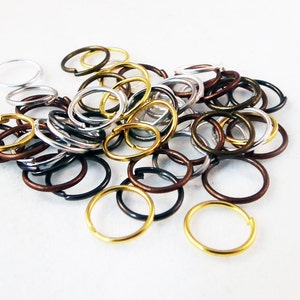 Può includere: Una collezione di anelli di giunzione in metallo di vari colori, tra cui argento, oro, bronzo e nero. Gli anelli sono rotondi e sparsi su una superficie bianca. Questi piccoli anelli sono spesso usati nella creazione di gioielli e nell'artigianato.
