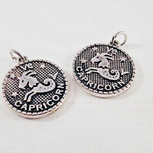 May include: Two silver-colored Capricorn zodiac charms. Each charm features a detailed image of a goat and the word "CAPRICORN" in a circular design. The charms have a small loop at the top for attaching to a chain or bracelet.