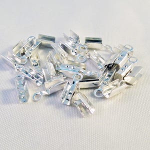 Puede incluir: Un montón de terminales de crimpado de metal plateado para la fabricación de joyas. Los terminales tienen un pequeño bucle en un extremo y una forma rectangular plana.