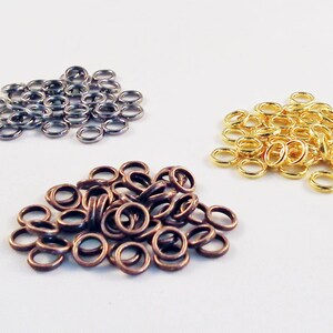 Op de afbeelding: Een stapel zilveren springringen, een stapel bronzen springringen en een stapel gouden springringen. Dit zijn kleine metalen ringen die worden gebruikt voor het maken van sieraden.