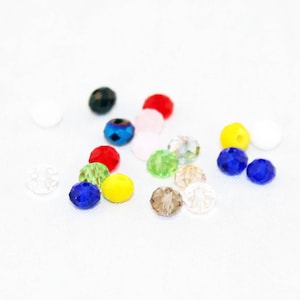 May include: Assortment of colorful faceted glass beads in various colors, including red, blue, green, yellow, and clear. The beads are scattered on a white surface.
