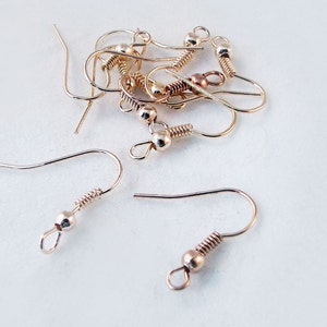 Peut inclure: Gros plan sur un groupe de crochets de boucles d'oreilles dorés. Les crochets sont en métal et ont une petite perle ronde au sommet. Les crochets sont disposés en pile sur une surface blanche.