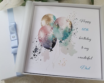 May include: A white card with a blue ribbon and a blue bow. The card has a watercolor illustration of three balloons in shades of teal, pink, and gold. The text on the card reads "Happy 60th birthday to my wonderful Dad."