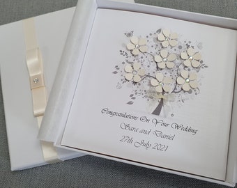 Könnte beinhalten: Eine weiße Karte mit einem grauen Baumdesign und weißen Blumen mit Strasssteinen. Die Karte befindet sich in einer weißen Schachtel mit einem weißen Satinband. Der Text auf der Karte lautet "Herzlichen Glückwunsch zur Hochzeit, Sara und Daniel, 27. Juli 2021".