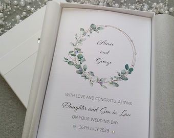 May include: White card with a green and gold eucalyptus wreath with rhinestones. The card says "Aimee and George" inside the wreath. The text below the wreath says "With love and congratulations Daughter and Son in Law on your wedding day 16th July 2023".