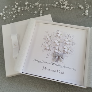 May include: A white card with a silver ribbon and a silver glitter butterfly on a white background. The card says "Happy Diamond Wedding Anniversary Mum and Dad".