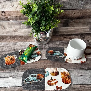 May include: Oval fabric coasters with fall-themed designs, including pumpkins, sunflowers, and vintage trucks. One coaster has a white mug with a brown handle. A green granola bar rests on a coaster. A silver vase holds a green plant.