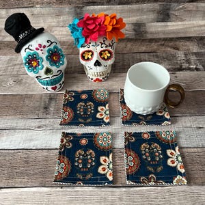 May include: A Day of the Dead themed collection featuring skull figurines and coasters. The skull figurines are white with colorful floral and decorative accents. The coasters are navy blue with a skull and floral pattern. A white mug with a gold handle is also present.