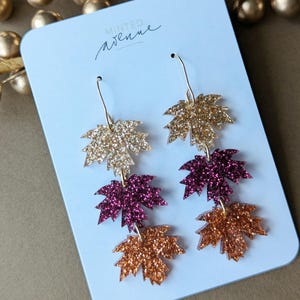 May include: Glittery leaf earrings in gold, burgundy, and copper colors. The earrings are hanging on a light blue card with the text "Minted Avenue" in cursive. The earrings are a fall-themed accessory.