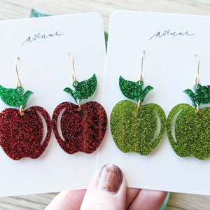 May include: A pair of dangle earrings featuring red and green glitter apple shapes with green glitter leaves. The earrings are on white cards with the text "Minted Avenue" printed on them.