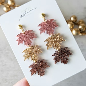 May include: A pair of dangle earrings featuring three layered maple leaf charms. The leaves are made of resin and have a glitter finish. The top leaf is pink, the middle leaf is gold, and the bottom leaf is brown. The earrings are on a white card with the text "Minted Avenue".