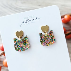 May include: A pair of gold glitter cat earrings with a gold heart charm. The earrings are on a white card with the text "Minted Avenue".