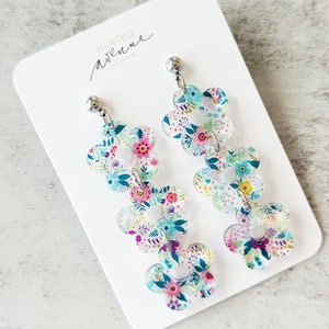 May include: Floral dangle earrings with a layered flower design. Each earring features three flower-shaped layers with a colorful floral pattern in shades of blue, pink, and white. The earrings are attached to a silver post. The card reads "Minted Avenue."