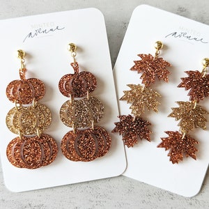 May include: Two pairs of dangle earrings. One pair features three stacked glittery pumpkins in shades of gold and copper. The other pair has three glittery maple leaves in similar colors. Both pairs have gold-colored ear wires.