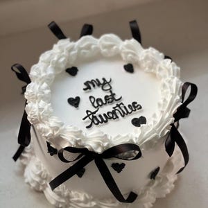 Pode incluir: Um bolo redondo branco com decorações pretas. O bolo é adornado com corações pretos e fitas pretas. As palavras "my last twenties" estão escritas em preto no topo.