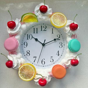 Puede incluir: Un reloj de pared decorativo con esfera blanca y números negros. El reloj está rodeado por una corona de glaseado blanco, cerezas rojas, rodajas de limón, rodajas de lima y macarons de colores. Las manecillas del reloj señalan las 10:10.