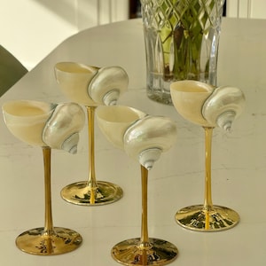 May include: Four decorative wine glasses. Each glass has a natural seashell bowl, a gold-coloured stem, and a circular base. The glasses are displayed on a white surface, with a crystal vase in the background.