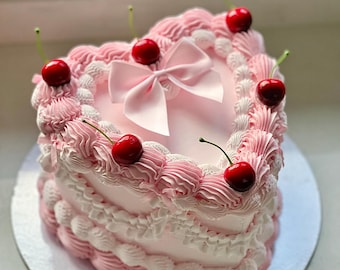 Può includere: Una torta a forma di cuore a due piani decorata con glassa rosa e bianca. La torta è sormontata da un fiocco rosa e da diverse ciliegie rosse.