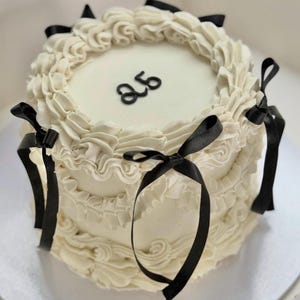 Könnte beinhalten: Ein runder, zweistöckiger Kuchen mit weißem Zuckerguss und schwarzen Schleifen. Auf der Oberseite des Kuchens steht die Zahl "25" in Schwarz. Der Kuchen ist mit gerüschtem Zuckerguss und schwarzen Schleifen verziert.